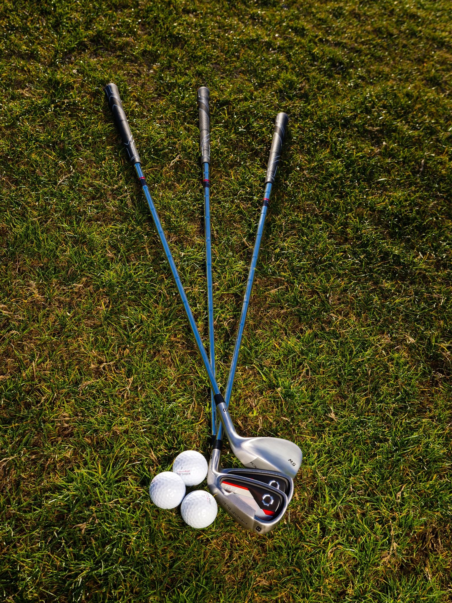 Three golf clubs and balls neatly arranged on green grass under daylight.